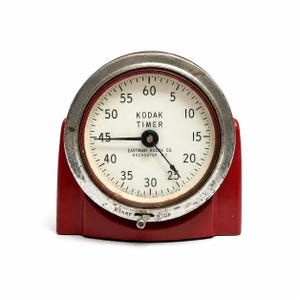 May include: A vintage Kodak Timer with a round white face, black numerals from 5 to 60, and two black hands. The device features a metallic silver bezel and a red base with a small metal lever at the bottom labeled START and STOP.