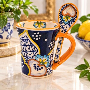 May include: A ceramic mug featuring a deep blue base with intricate white, yellow, and light blue floral and geometric patterns. The mug includes an integrated orange handle and a matching spoon that rests in a slot on the side of the mug.
