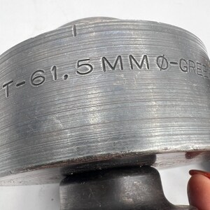 Pode incluir: Close-up de um componente met&aacute;lico industrial com o texto "T-61.5MM &Oslash;-GREEN" gravado em sua superf&iacute;cie. O objeto tem uma forma cil&iacute;ndrica com uma textura estriada e uma apar&ecirc;ncia escura e envelhecida. O componente parece ser feito de um metal dur&aacute;vel.