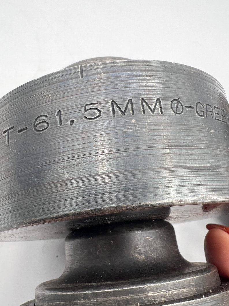 Pode incluir: Close-up de um componente met&aacute;lico industrial com o texto "T-61.5MM &Oslash;-GREEN" gravado em sua superf&iacute;cie. O objeto tem uma forma cil&iacute;ndrica com uma textura estriada e uma apar&ecirc;ncia escura e envelhecida. O componente parece ser feito de um metal dur&aacute;vel.