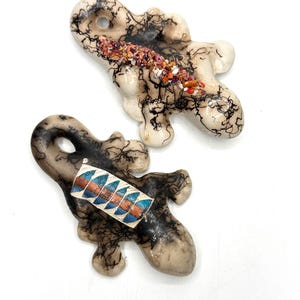 May include: Two irregular, lizard-shaped ceramic ornaments featuring a black horsehair-fired pattern on a cream-colored base. One ornament has a central strip of crushed orange and red stones, while the other features a rectangular inlay with blue and copper geometric patterns.