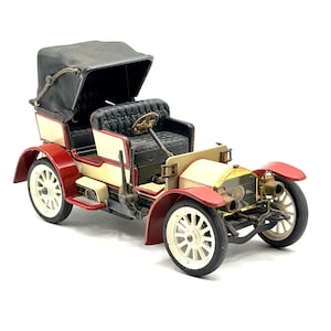 May include: A vintage model car in red, cream, and black. The car features a black convertible top, cream-colored body panels, and red fenders. The wheels are white with a spoke design. The car has a gold-colored front grill.