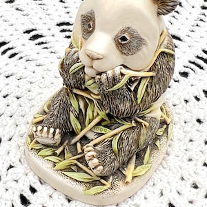 May include: A detailed figurine of a panda bear seated and eating bamboo. The panda has a cream-colored face and body with black fur around the eyes, ears, and shoulders. The bamboo is green and yellow. The base is cream-colored with the number "1000" visible.