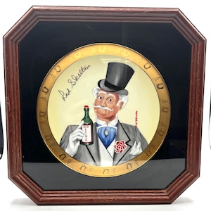 May include: Framed artwork featuring a painted portrait of Red Skelton holding a green bottle. The artwork is in a dark wood octagonal frame with a black background. The portrait has a gold border with decorative elements.