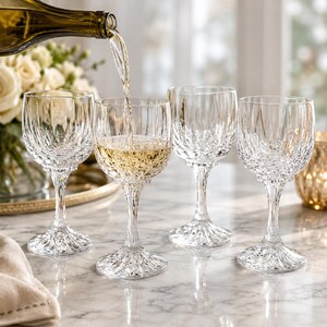 May include: Four clear crystal wine glasses with intricate diamond-cut patterns. One glass is being filled with white wine from a bottle. The glasses are on a marble surface, with a floral arrangement and a candle holder in the background.
