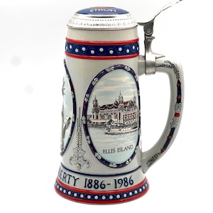 May include: A ceramic beer stein with a hinged lid. The stein features images of the Statue of Liberty and Ellis Island, with the text "LIBERTY 1886-1986". The lid is blue with the word "STROH". The stein has a silver handle and trim.