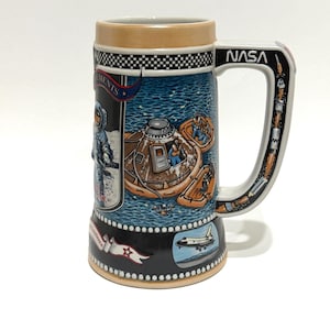 May include: A ceramic beer stein with NASA branding and space-themed illustrations. The mug features images of astronauts, spacecraft, and the NASA logo. The handle is white with a black and gray design. The top and bottom rims are black with a white checkered pattern.