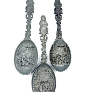 May include: Three antique silver-toned spoons with detailed relief carvings. Each spoon features a different scene of people and buildings, with a decorative figure at the handle's top. The spoons are displayed against a white background.