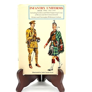 May include: A book titled "Infantry Uniforms Book Two 1855-1939" featuring illustrations of British military uniforms. The cover displays two soldiers in detailed period attire, one in khaki and the other in a traditional Scottish kilt. The book is displayed on a wooden stand.