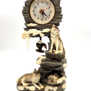 May include: A decorative clock featuring a wolf theme. The clock has a round face with black numbers and gold trim. The base is a rock formation with a howling wolf, an eagle, and two other wolves. The clock is approximately 10 inches tall.