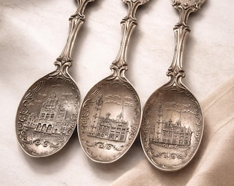 May include: Three antique silver spoons with detailed engravings of buildings on the bowls. Each spoon has a handle with the year "1989" at the top. The spoons are displayed on a neutral-toned surface with a beige cloth.