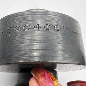 Pode incluir: Close-up de um objeto de metal cinza escuro com o n&uacute;mero "-5004063-" gravado em sua superf&iacute;cie. O objeto tem formato cil&iacute;ndrico com ranhuras horizontais. A parte inferior &eacute; um tom de cinza mais escuro. O fundo &eacute; branco.