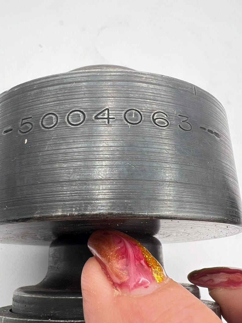 Pode incluir: Close-up de um objeto de metal cinza escuro com o n&uacute;mero "-5004063-" gravado em sua superf&iacute;cie. O objeto tem formato cil&iacute;ndrico com ranhuras horizontais. A parte inferior &eacute; um tom de cinza mais escuro. O fundo &eacute; branco.