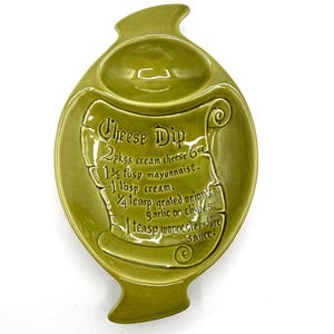 May include: A green ceramic cheese dip serving dish. The dish has a unique shape with a central scroll design that includes a recipe for cheese dip. The dish has a small bowl at the top. The text on the scroll reads "Cheese Dip".