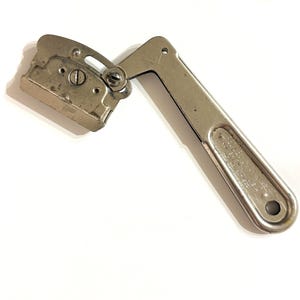 May include: An antique silver-toned can opener with a hinged design. The can opener has a rectangular cutting head and a long handle with a hole at the end. The handle has text on it.