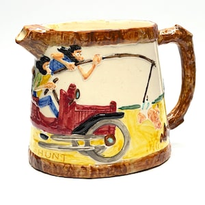 May include: A vintage ceramic jug with a brown handle and rim. The jug depicts a scene of a red car with two figures, and the word "HUNT" is printed on the base. The background is cream-coloured with a yellow and green landscape.