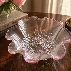 May include: A clear glass bowl with a ruffled edge and a pink tint. The bowl features a floral design in the center and is sitting on a dark wooden surface. The text "A BEAUTIFUL HOME" is visible in the background.