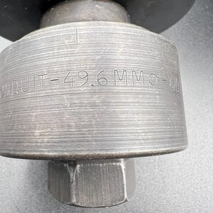 Pode incluir: Close-up de uma ferramenta de metal com o texto "CONDUIT-49.6 MM &Oslash;". A ferramenta tem um corpo cil&iacute;ndrico com ranhuras horizontais e uma base hexagonal. A parte superior &eacute; um disco plano grande.