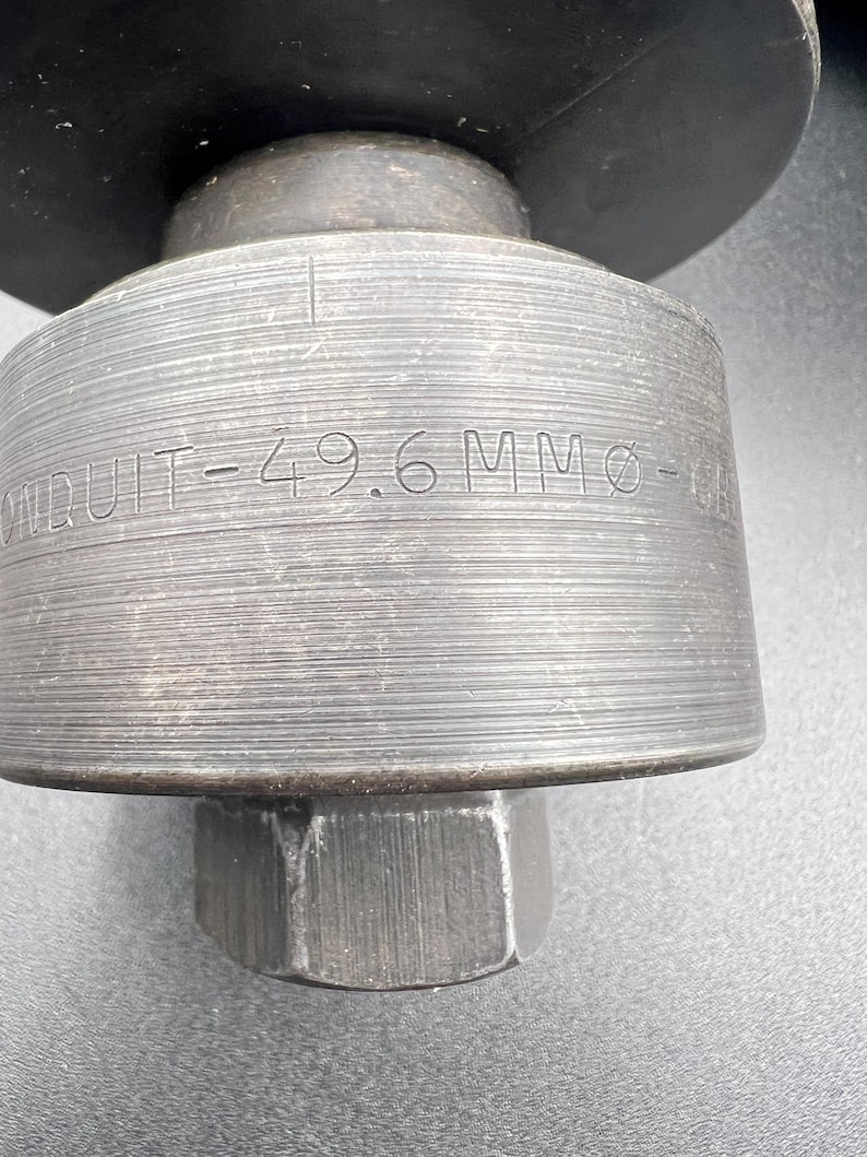 Pode incluir: Close-up de uma ferramenta de metal com o texto "CONDUIT-49.6 MM &Oslash;". A ferramenta tem um corpo cil&iacute;ndrico com ranhuras horizontais e uma base hexagonal. A parte superior &eacute; um disco plano grande.