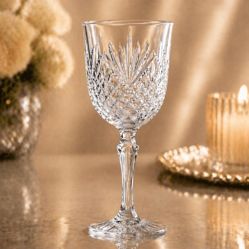May include: A clear crystal wine glass with intricate cut patterns. The glass features a detailed design on the bowl and stem, reflecting light. The background includes a blurred candle and decorative elements, suggesting a formal setting.