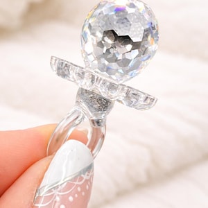 May include: A clear, crystal-like pacifier with a faceted, spherical top. The pacifier has a round handle and a flat, petal-shaped guard. The object is held against a soft, white background.