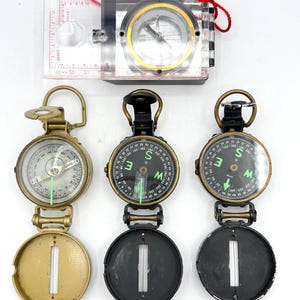 May include: A collection of compasses, including a clear plastic baseplate compass with a red lanyard and several vintage-style compasses with black and gold casings. The compasses have rotating dials and luminous needles.