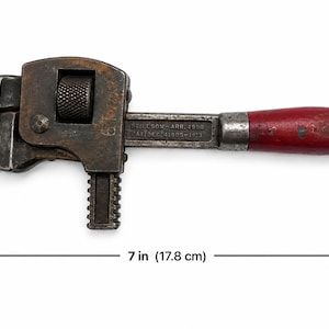 May include: An antique Stillson pipe wrench, 7 inches (17.8 cm) long, with a rusty, dark metal finish. It features a red wooden handle and adjustable jaws. The wrench is designed for gripping and turning pipes.