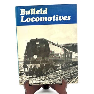 May include: A vintage book titled "Bulleid Locomotives" with a blue and white cover. The book features a black and white image of a steam train on the front. The book is displayed on a brown wooden stand.
