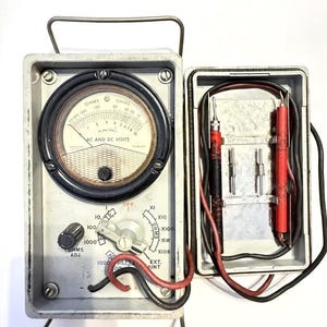 May include: Vintage gray multimeter with a black dial displaying OHMS and AC and DC VOLTS. The meter has a needle indicator, a knob, and various settings. The open case reveals probes and additional components.
