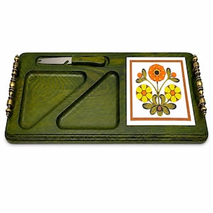 May include: A rectangular green wooden serving board featuring two triangular recessed sections, a metal cheese knife with a matching green handle, and a square ceramic tile inlay decorated with a floral pattern of orange and yellow flowers.
