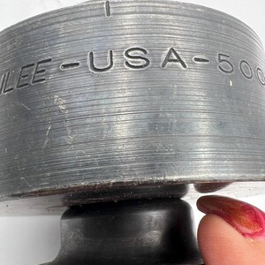 Pode incluir: Close-up de um objeto de metal cinza escuro com o texto "LEE - USA - 500" gravado na lateral. O objeto tem uma textura estriada e uma forma circular. Um dedo com esmalte vermelho est&aacute; tocando o objeto.