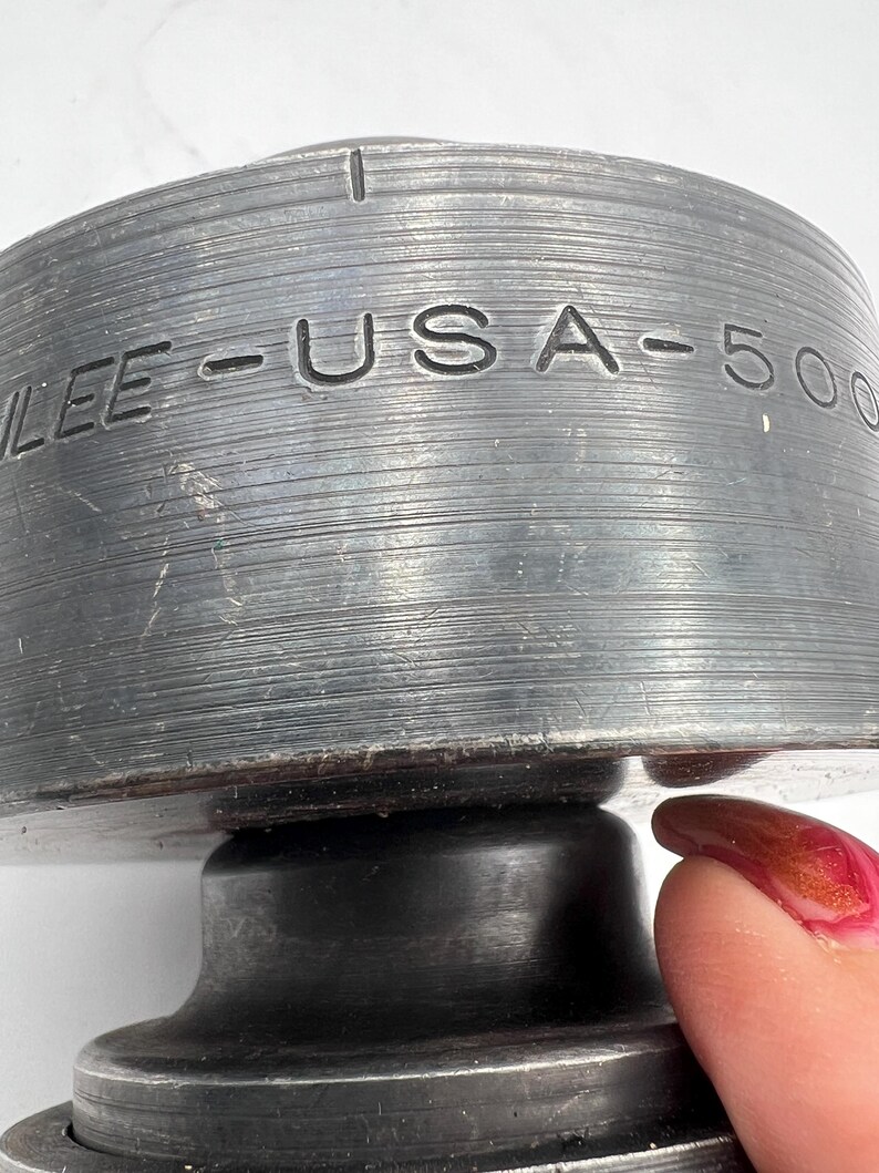 Pode incluir: Close-up de um objeto de metal cinza escuro com o texto "LEE - USA - 500" gravado na lateral. O objeto tem uma textura estriada e uma forma circular. Um dedo com esmalte vermelho est&aacute; tocando o objeto.