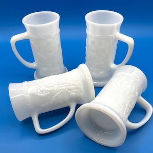May include: Four white glass beer steins with handles, arranged on a blue surface. The steins have a detailed embossed design and a classic shape. The steins are made of a milky white glass.