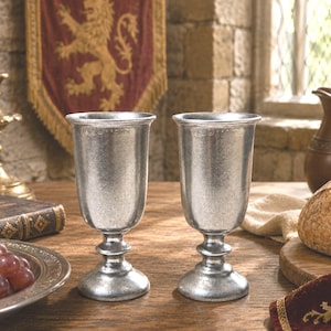 May include: Two silver-colored goblets with detailed bases and rims sit on a wooden table. A lit candle in a gold candlestick, a book, and a plate of grapes are also on the table. A loaf of bread and a pitcher are in the background.