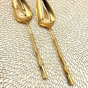 May include: Two gold-colored serving spoons with bamboo-style handles. One spoon has a slotted design. The spoons are on a gold-colored, textured surface. The spoons are likely for serving food.