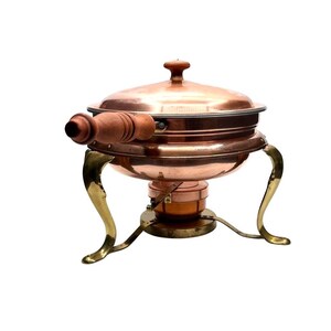 May include: A copper chafing dish with a polished finish, featuring a round lid with a small knob handle, a side handle made of turned wood, and three curved brass legs. A small copper burner unit is positioned centrally beneath the main bowl.