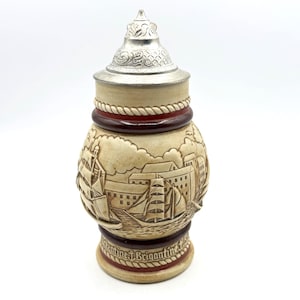 May include: A beige ceramic stein with a silver lid. The stein features a detailed relief of sailing ships and buildings. A dark red band with a rope design encircles the middle. The base has text that reads "Valentine J. Brigantine 1986".