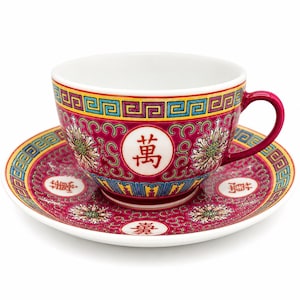 May include: A magenta porcelain teacup and saucer set featuring intricate floral patterns, a yellow and blue geometric border, and white circular medallions containing red Chinese characters. The cup has a matching magenta handle and white interior.