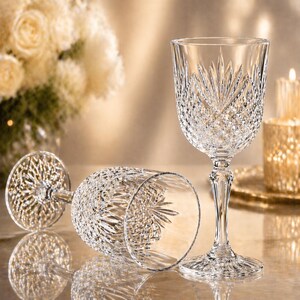 May include: Two clear crystal wine glasses with intricate cut glass patterns. One glass stands upright, while the other lies on its side. The glasses are on a reflective surface, with a soft, blurred background featuring flowers and a candle.