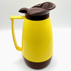 May include: A vintage yellow and brown insulated pitcher with a handle and a lid. The pitcher has a yellow body, a brown base, and a brown lid. The handle is yellow. The pitcher is designed to keep liquids hot or cold.
