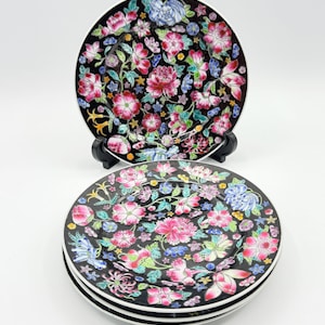 May include: A set of decorative plates with a black background and a vibrant floral pattern. The plates feature pink, blue, and white flowers with green leaves. The plates have a white rim and are stacked on top of each other.