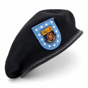 May include: A black wool beret featuring a leather trim along the edge and a blue embroidered patch. The patch displays a shield with a bear emblem, surrounded by white stars, and includes the Latin text OMNE VIR TIGRIS at the bottom.