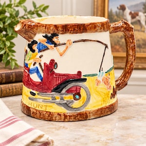 May include: A ceramic pitcher featuring a relief design of two figures in a red vintage car, with one figure holding a fishing rod. The rim and handle are textured to resemble wood grain. The word HUNT is printed in yellow near the base.