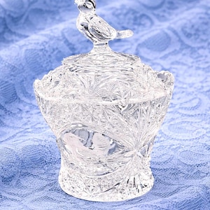 May include: A clear, decorative glass jar with a lid, featuring a bird-shaped handle. The jar has a detailed, cut-glass design with floral and starburst patterns. The jar is sitting on a blue lace fabric.