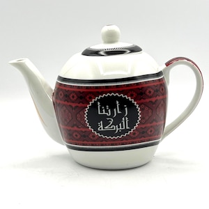 May include: White ceramic teapot with a red and black patterned design around the body. The teapot has a white lid and handle with a black band around the top. The Arabic text is displayed on the front of the teapot.