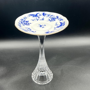 May include: A clear glass pedestal with a decorative base supports a white saucer. The saucer features a blue floral design and a gold rim. The pedestal is approximately 8 inches tall.