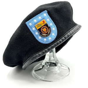 May include: Black beret with a leather trim and a patch featuring a shield, stars, and the words "OMNE VIR TIGRIS". The beret is displayed on a clear glass stand.