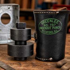 Pode incluir: Uma ferramenta de pun&ccedil;&atilde;o preta com uma porca sextavada e uma bolsa de couro. A ferramenta &eacute; rotulada como "GREENLEE NO. 737 KNOCKOUT PUNCH ROCKFORD, ILL." Projetada para trabalhos de condu&iacute;tes el&eacute;tricos. A bolsa &eacute; preta com um r&oacute;tulo verde.
