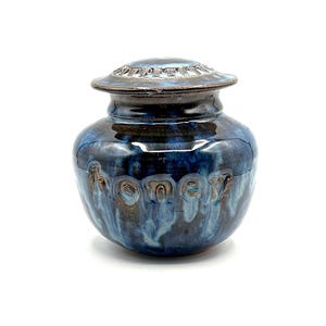 May include: A ceramic honey pot with a lid. The pot is a deep blue with lighter blue and brown streaks, and the word "honey" is embossed on the front. The lid has a decorative rim.