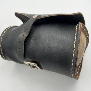 Pode incluir: Um estojo de couro preto com aba e fecho de correia. O estojo cil&iacute;ndrico apresenta costuras brancas e uma fivela prateada. &Eacute; projetado para guardar ferramentas ou objetos pequenos. Aproximadamente 20 cm de comprimento.